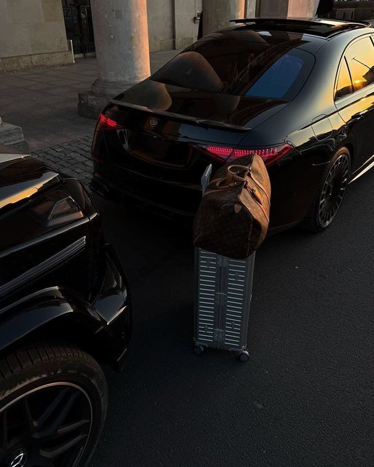 A black luxury sedan parked on a cobblestone street at sunset with a Louis Vuitton monogram duffel bag placed on a silver rolling suitcase next to it.