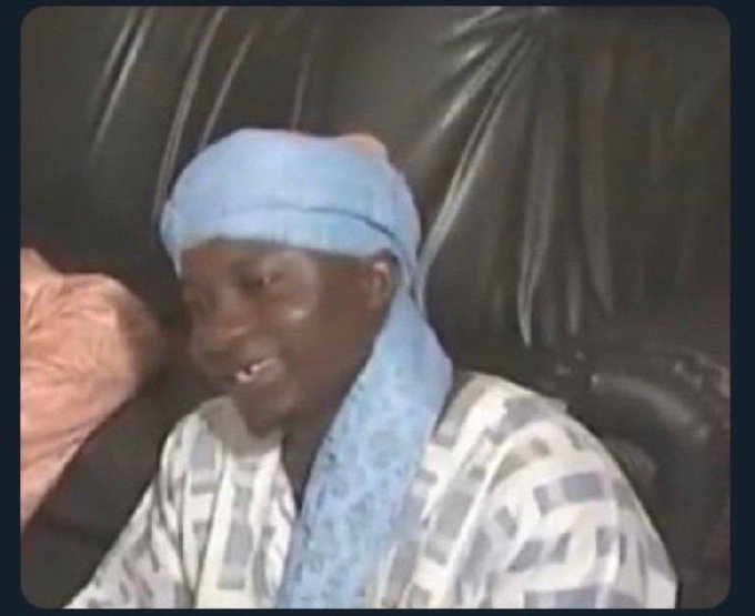 An older video still of a person wearing a blue head wrap and patterned robe sitting on a dark leather couch.