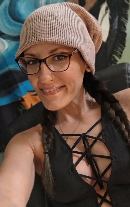 A selfie of a smiling woman with braided pigtails wearing a pink beanie, glasses, and a black strappy top.