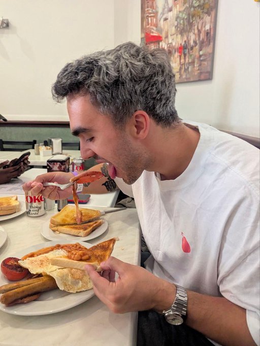A man with curly salt-and-pepper hair is enthusiastically eating bacon with his hands while seated at a diner table featuring plates of toast, sausages, and eggs.