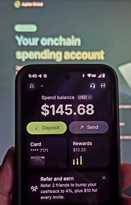 A hand holding a smartphone displaying an onchain spending account app with a spend balance of $145.68, deposit and send options, and a 'Refer and earn' banner.