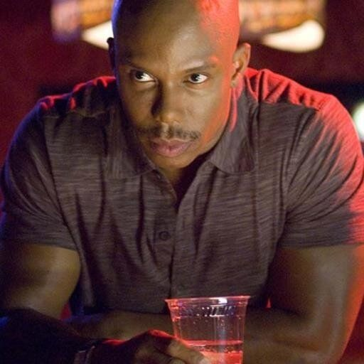 A serious-looking man in a dark short-sleeved button-up shirt sits at a bar holding a glass of liquid, illuminated by dramatic red and purple lighting.