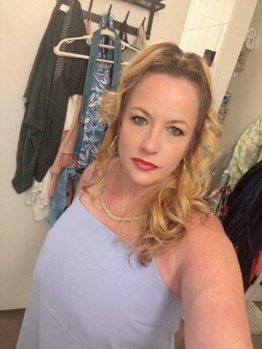 A woman with blonde curly hair and red lipstick poses in a light blue tank top, accessorized with layered gold necklaces, in front of a closet rack filled with clothes. The selfie complements her "Gm Sunday Funday" post, capturing a relaxed, ready-to-go vibe perfect for a fun Sunday outing.