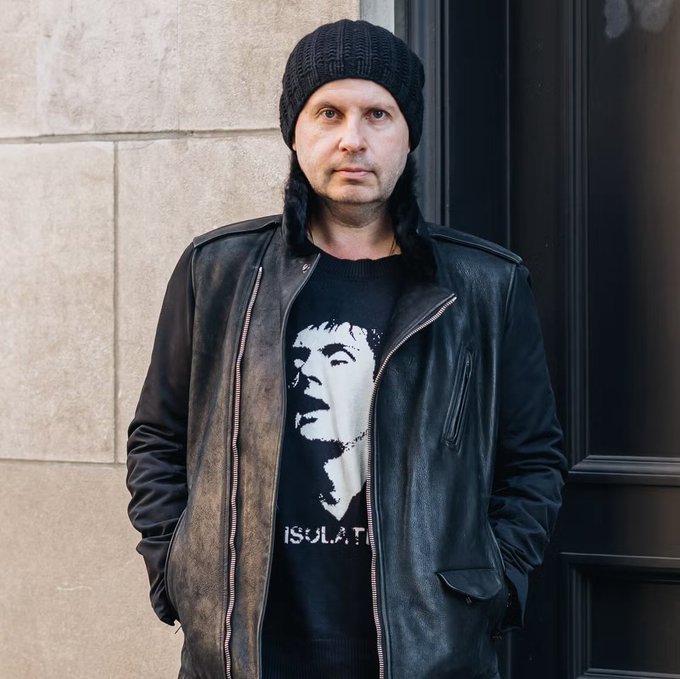A man with long dark hair wearing a black knit beanie, a distressed leather jacket, and a black t-shirt featuring a graphic and the word 'ISOLATI' stands outdoors against a stone wall.