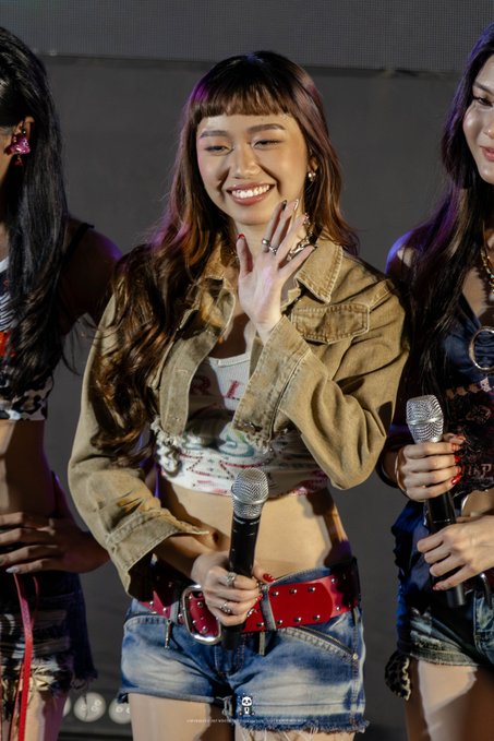 A young woman with bangs and long hair smiles and waves while holding a microphone on stage, wearing a cropped denim jacket, a graphic crop top, and denim shorts with a red belt.