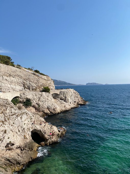 A scenic coastal view features rugged limestone cliffs meeting clear turquoise Mediterranean waters, with swimmers enjoying the sea and a distant landmass visible across the bay. The image pairs with the "GM to everyone" message by offering a bright, uplifting morning landscape to greet viewers with a cheerful start to the day.