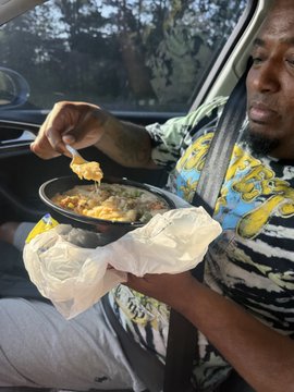 A man in a colorful patterned shirt sits in a car, eating a piece of fried food from a black takeout bowl.