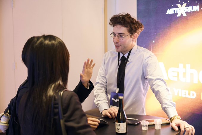 Photo of a young man wearing a 'SPEAKER' badge and glasses talking to a woman at what appears to be an Aethirum event booth, with sake and cups on the table.