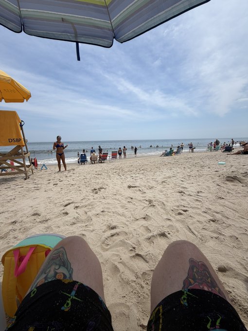 A first-person view from a beach chair under a blue and white umbrella shows a person's legs extended, looking out at a sandy beach with a lifeguard stand, ocean waves, and beachgoers.