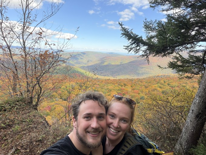 A smiling couple takes a selfie atop a scenic overlook, framed by a stunning vista of rolling mountains blanketed in vibrant fall foliage under a bright blue sky. "gm Legends" captures the upbeat, adventurous spirit of starting a fresh fall day with a loved one in the great outdoors.