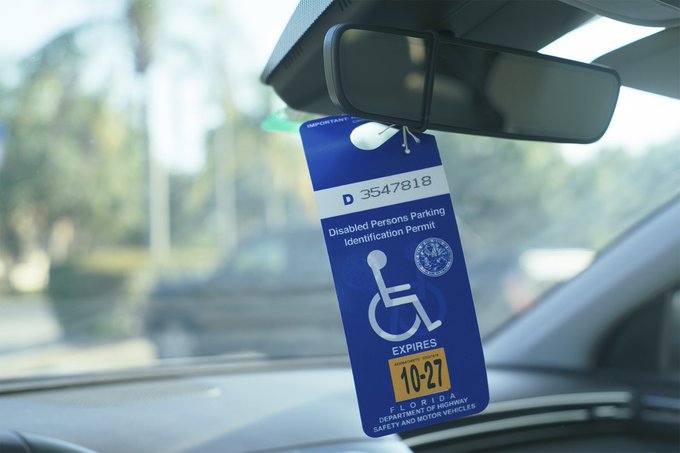 A blue 'Disabled Persons Parking Identification Permit' hangs from a car's rearview mirror, with the number 'D 3547818' and an expiration date of '10-27', issued by Florida Department of Highway Safety and Motor Vehicles.