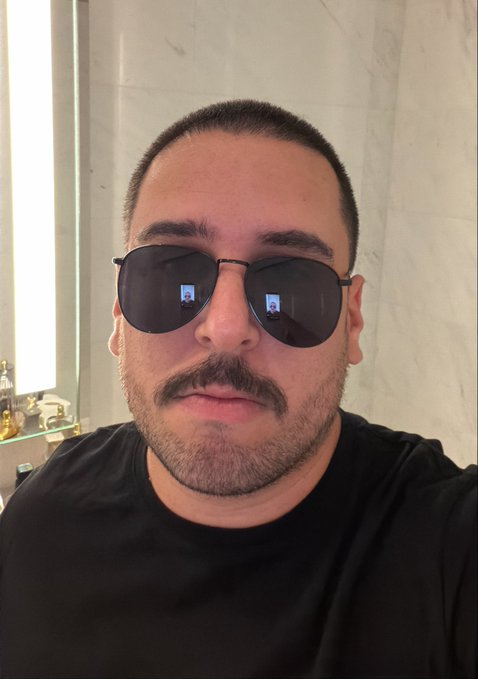 A man wearing aviator sunglasses takes a selfie in a marble-walled bathroom, with phone screen reflections visible in his lenses. The "trading cop" persona fits the serious, watchful expression behind the shades, reinforcing the post's message about policing bad trading habits.