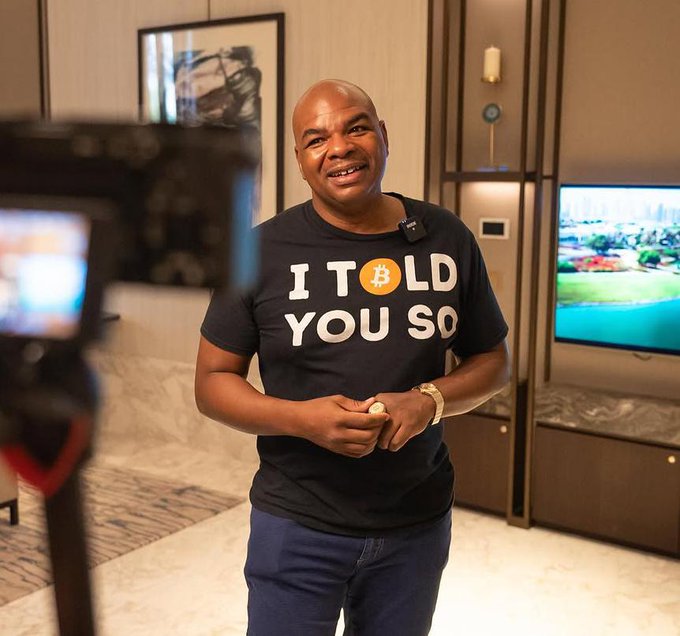 A man wearing a black t-shirt that reads 'I TOLD YOU SO' with a Bitcoin 'B' logo instead of the 'O', standing in what appears to be a hotel room.
