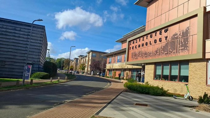 Street view of a university campus featuring a modern building labeled 'INSTITUTE OF TECHNOLOGY' with a decorative facade, alongside other buildings and a green scooter parked nearby.