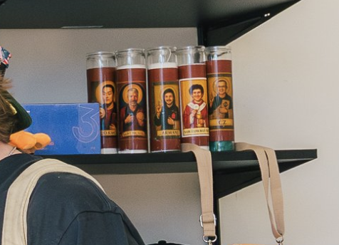 Five saint candles labeled "ST. JEROME," "ST. MAXIMILIAN KOLBE," "ST. JOHN," "ST. JOHN," and "ST. FRANCIS" are lined up on a black shelf next to a blue sign with the number "3," with a tan bag and backpack visible below.
