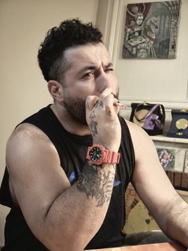 The image features a man with tattoos sitting at a table, holding a cigarette and looking down. The background includes a painting of a woman's face and a clock. The post reads "gm. artist. v art: Lullaby of Moon Lake - 22.19 cm 0.2 Ξ".