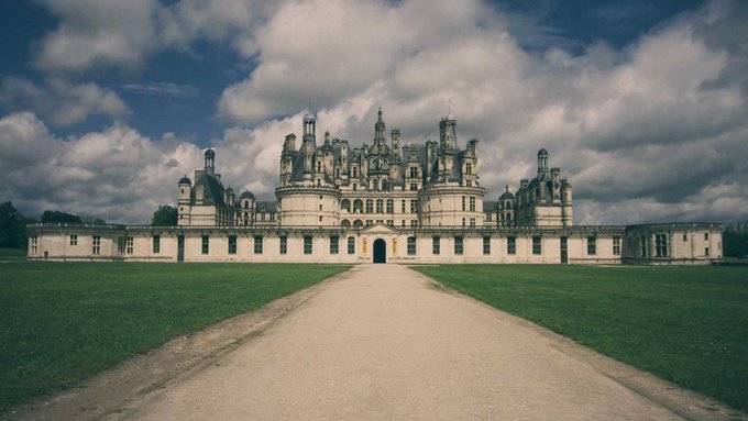 The image features a large white castle or building situated in a grassy area with a dirt road leading up to it, giving an impression of grandeur and majesty. The castle appears to be old and possibly abandoned, adding to its historical charm. The surrounding landscape includes trees and other buildings, creating a picturesque scene that complements the castle's architectural beauty.