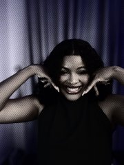 A smiling woman with dark hair poses with her hands near her face against a dark curtain background.