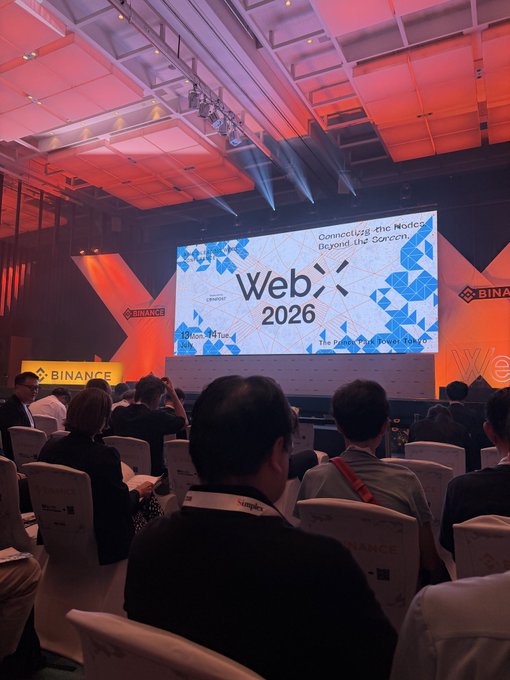 Attendees sit facing a stage at WebX 2026 with a large screen displaying "WebX 2026" and "Connecting the World, Beyond the Sorrens" beneath Binance-branded signage and red-lit ceiling beams, matching the post's observation of Binance's dominant presence as the flagship sponsor ahead of Prime Minister Sanae Takaichi's appearance.
