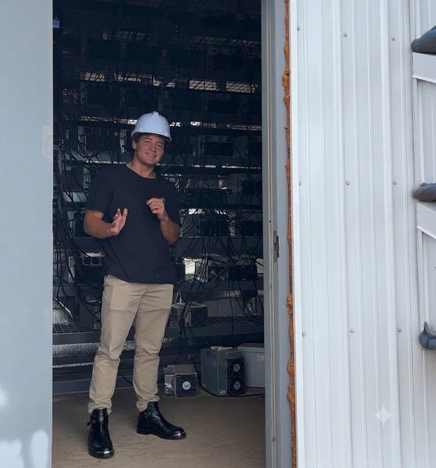 The image features a man wearing a white hard hat, standing inside an industrial setting with a large number of black boxes surrounding him. He appears to be engaged in some sort of work or activity related to the stack of real bitcoin. The man is positioned near a door and seems to be making a gesture towards it.