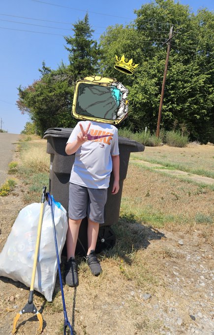 A child wearing a cartoon TV head with a drawn-on crown and white t-shirt makes a peace sign while standing next to a black trash can outdoors, holding a grabber tool.