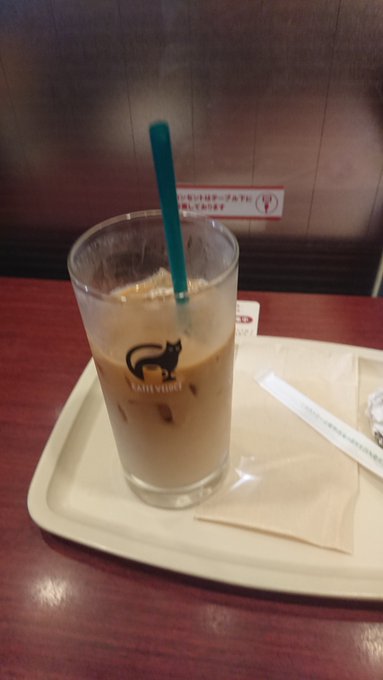 A glass of iced coffee with a blue straw and a cat logo sits on a white tray with napkins and a receipt on a wooden table.
