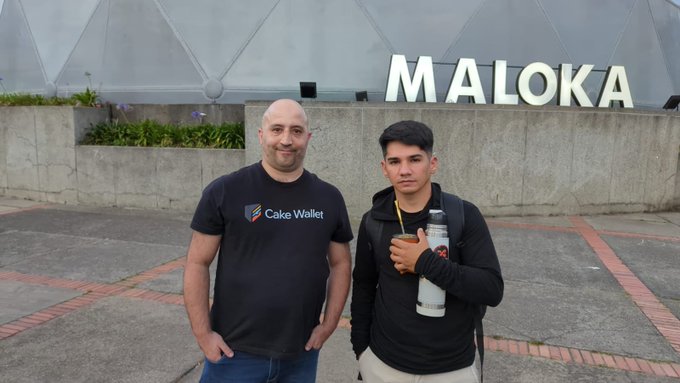 Two men posing for a photo in front of a concrete wall with large white letters spelling 'MALOKA'; the man on the left wears a Cake Wallet branded t-shirt, while the man on the right holds a water bottle and wears a black hoodie.
