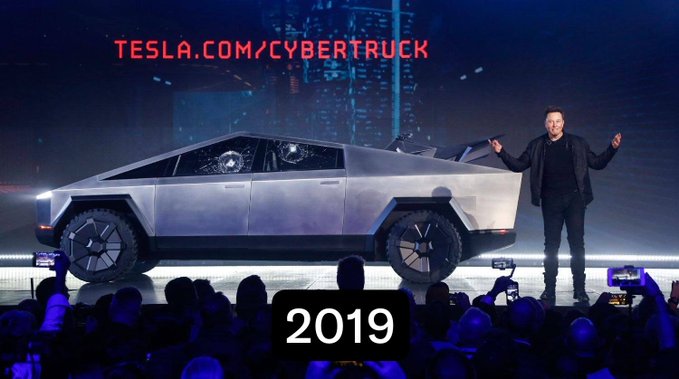 Tesla's Cybertruck is unveiled on stage with Elon Musk presenting, with 'TESLA.COM/CYBERTRUCK' displayed at the top and '2019' labeled below.