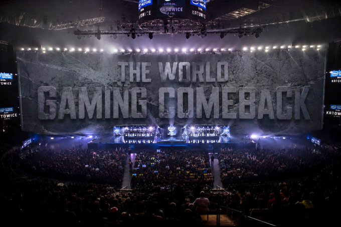 A massive esports arena filled with spectators, featuring a large stone-textured screen displaying the text 'THE WORLD GAMING COMEBACK' under dramatic stage lighting.
