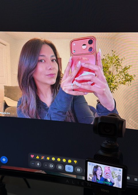 A computer monitor displays a woman with long hair taking a mirror selfie with a pink phone, while a DJI Osmo camera in the foreground shows a live feed of the same image.