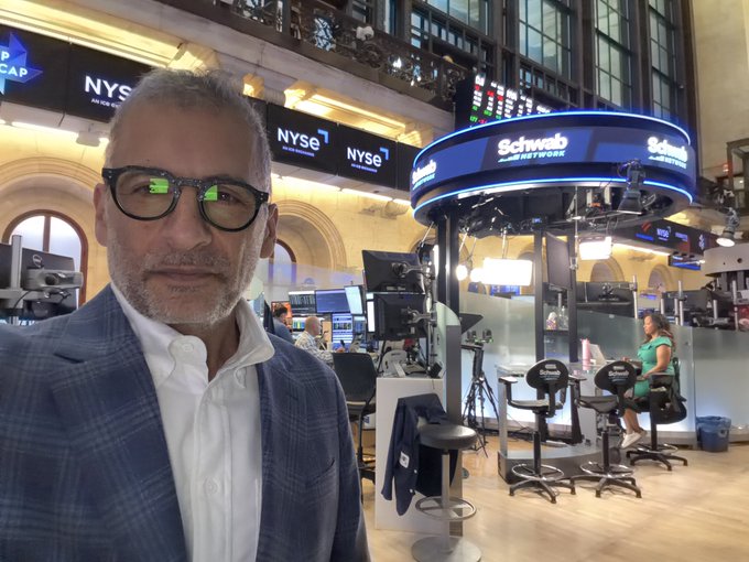A selfie of a man with glasses and a gray beard wearing a blue plaid blazer, taken at the New York Stock Exchange (NYSE) trading floor with the Schwab Network broadcast booth visible behind him.