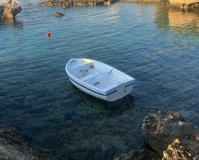 A small white rowboat with a blue stripe is anchored in clear turquoise water between rocky shores, with a small red buoy floating nearby. The image humorously contrasts the modest dinghy with the poster's wish to "trade in" their tiny vessel for a yacht.