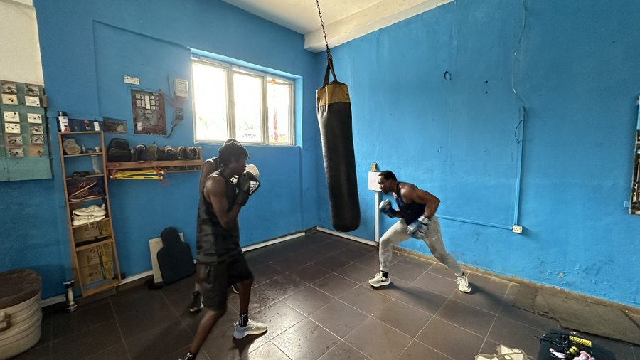 Two young men wearing boxing gloves training with a punching bag inside a blue-walled gym with tiled floors.
