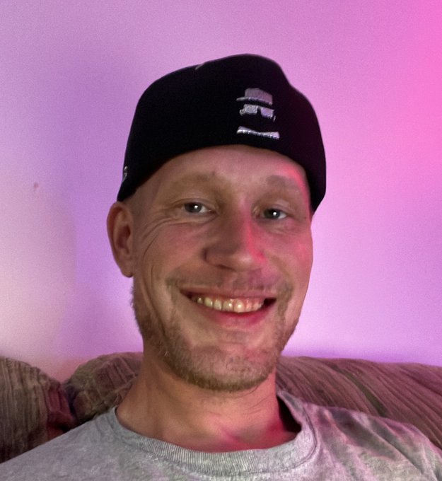 The image features a man wearing a black hat with a white logo, sitting comfortably on a couch against a pink wall. The man has a big smile on his face as he looks directly at the camera. The caption of the image reads "GM FAM hope everyone has a terrific Tezos Tuesday Or taco Tuesday...however you partake...make it a good one". This suggests that the man is enjoying himself and wants to share his positive experience with others, possibly in response to an event or promotion related to tacos on Tezos Tuesday.