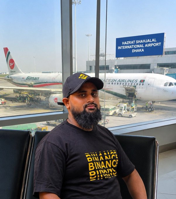 A man wearing a black "Binance" t-shirt sits at Hazrat Shahjalal International Airport in Dhaka with a Biman Bangladesh Airlines plane visible through the window behind him, illustrating his travels as a Binance Affiliate celebrating the platform's 9th Anniversary.
