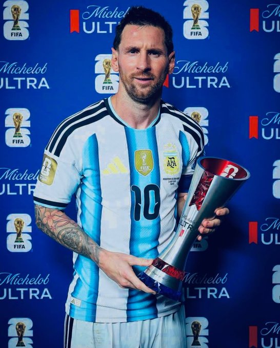 Lionel Messi wearing the Argentina #10 jersey, holding a silver and red trophy in front of a blue backdrop covered in FIFA World Cup and Michelob Ultra logos.