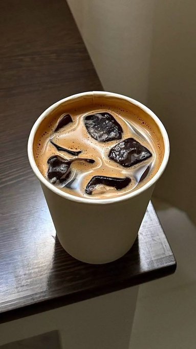 A paper cup filled with iced coffee containing several ice cubes sits on a dark wooden table.