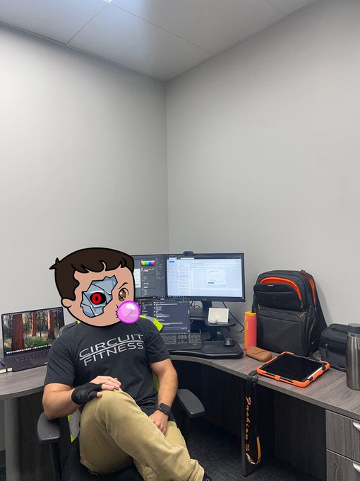 A man wearing a 'CIRCUIT FITNESS' t-shirt sits in a chair at a desk with multiple monitors, with a Terminator-style cyborg emoji sticker covering his face.