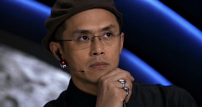 The image features a man wearing glasses, a hat, and holding a microphone with his hand over his mouth as if he's deep in thought or contemplation. The background of the image shows a blue wall with a planet visible behind it. There is also text that reads "The next monthly #Binance $LUNC burn is in 3 days! Binance has burned Terra Luna Classic every month since 202 and continues to support the Terra Luna Classic community. Thank you Binance for the support!". The man's position, along with his thoughtful expression, suggests he might be a creator or participant of the Terra Luna Classic series on Binance.