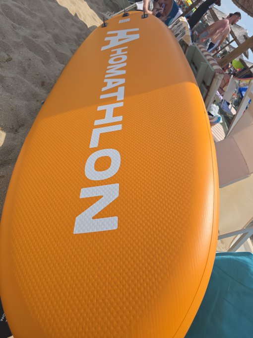 An orange stand-up paddleboard branded with "HAMATHLON" in large white letters, with people visible at a beach resort in the background.