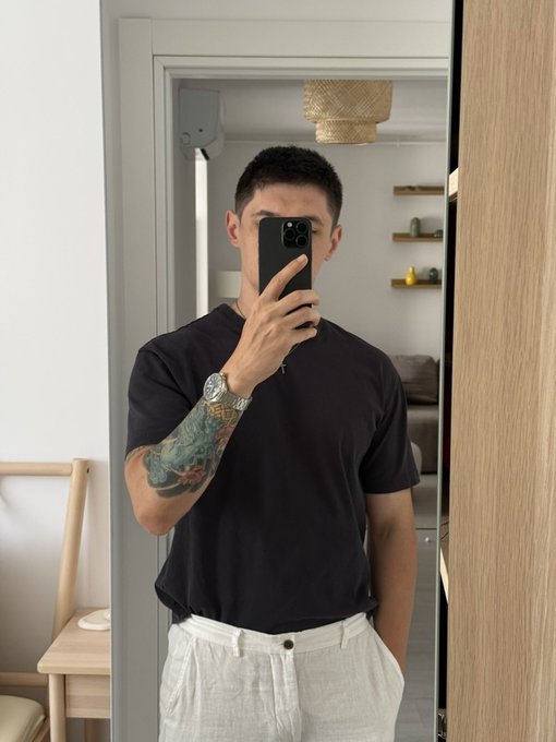A young man with a tattooed arm taking a mirror selfie with his phone, wearing a black t-shirt and light-colored pants in a modern interior.