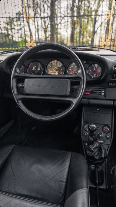 The interior view of a Porsche sports car showing the steering wheel with the Porsche logo, dashboard gauges, and manual gear shift.