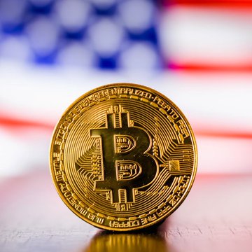 A gold Bitcoin coin stands upright against a blurred American flag backdrop, symbolizing the intersection of cryptocurrency and U.S. regulatory efforts referenced in the post about SEC Chair Paul Atkins pushing for the Bitcoin Clarity Act.