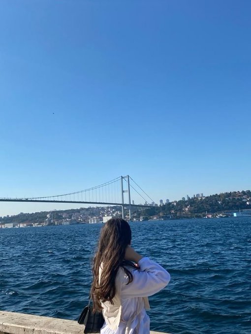 A woman with long dark hair stands facing the Bosphorus strait, looking toward a suspension bridge connecting two landmasses under a clear blue sky.