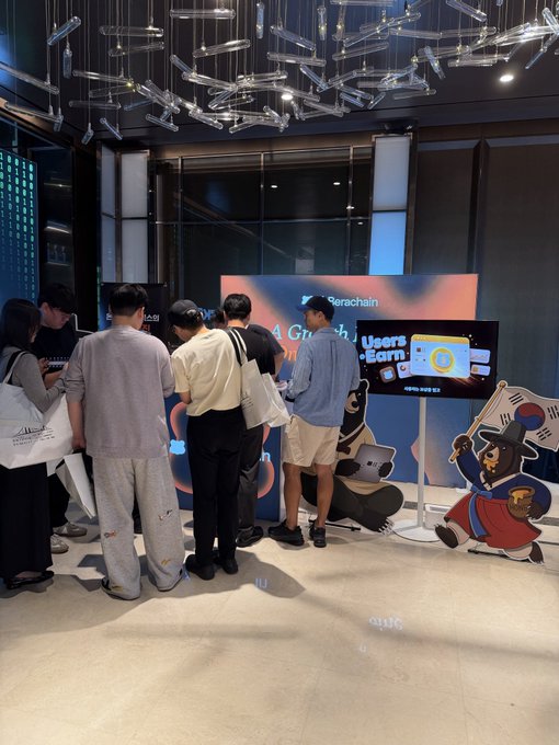 A group of attendees gathers around a Berachain booth featuring a digital display and a bear mascot dressed in traditional Korean attire with a South Korean flag, fitting the "Catch the Beras" theme at the Bridge Summit. The branded screen highlights Berachain and the slogan "A chain where Users Earn," with the event taking place in a sleek, modern venue adorned with hanging glass tube decor.