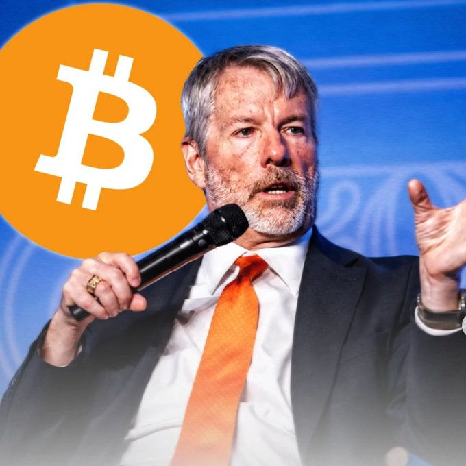 A man in a suit and orange tie speaking into a microphone, with a large orange Bitcoin logo superimposed in the upper left corner, likely referencing MicroStrategy's Michael Saylor.