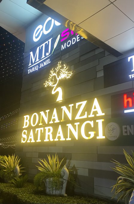 The image shows a large sign with white letters that reads "BONANZA SATRANGI" above some plants, possibly potted ones. The background of the photo features a building or store, which could be related to the sign's message about shopping for girls. There is also an advertisement posted on the wall, but it does not seem to be directly related to the "BONANZA SATRANGI" sign.