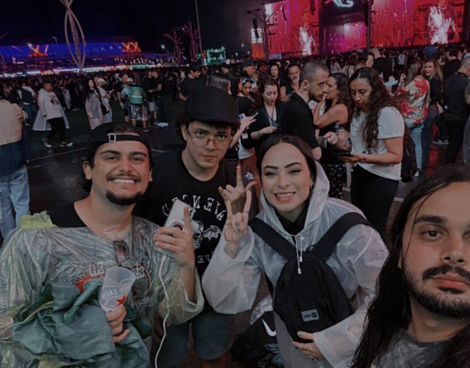 A selfie of four young people smiling and posing at a nighttime outdoor concert or festival, with a large crowd and a brightly lit red stage screen in the background.
