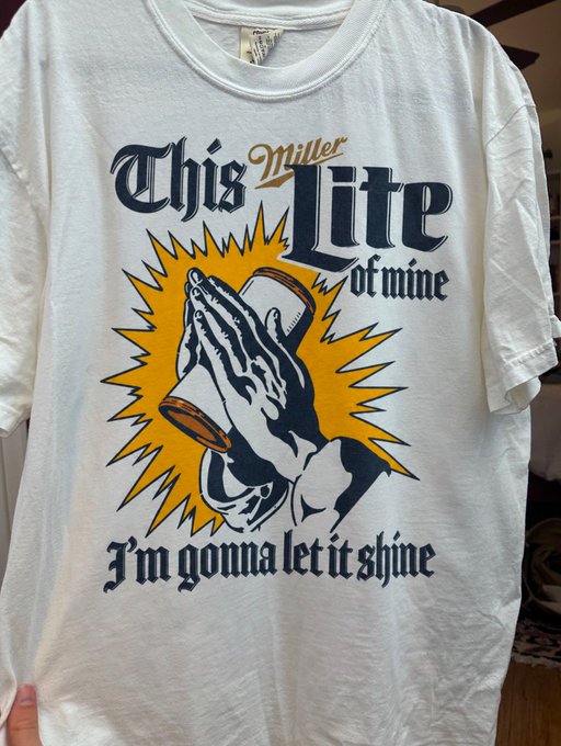 A white t-shirt featuring a graphic of praying hands holding a Miller Lite beer can with the text 'This Miller Lite of mine, I'm gonna let it shine' in stylized lettering.