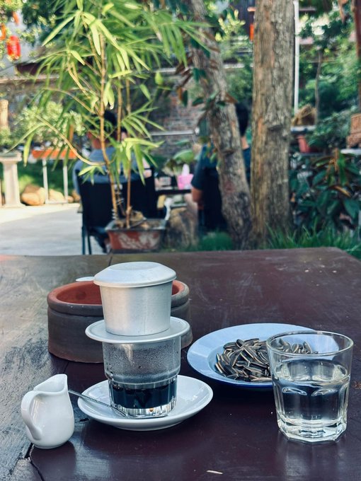A traditional Vietnamese coffee (cà phê phin) setup with a small white pitcher, a glass of water, and a plate of sunflower seeds sits on a wooden café table against a lush garden backdrop.

The image evokes the quiet morning café scene from the post—a moment of stillness and reflection over coffee, paired with the calm pause from trading BTC and ETH to simply enjoy the peaceful surroundings.